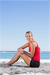 Athletic blonde sitting on sand smiling at camera on the beach