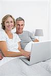 Middle aged couple using their laptop together in bed smiling at camera at home in bedroom
