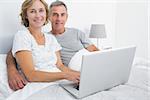 Relaxed couple using their laptop together in bed smiling at camera at home in bedroom