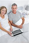 Couple using their laptop together in bed smiling at camera at home in bedroom