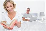 Woman reading book while husband is using laptop in bedroom at home