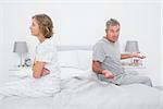 Couple sitting on different sides of bed having a dispute with husband gestruing at camera in bedroom at home