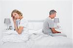 Annoyed couple sitting on different sides of bed having a dispute with woman looking at camera in bedroom at home