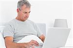 Grey haired man using his laptop in bed in bedroom at home