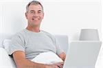 Happy grey haired man using his laptop in bed smiling at camera in bedroom at home