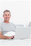 Cheerful grey haired man using his laptop in bed smiling at camera in bedroom at home