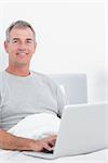 Smiling grey haired man using his laptop in bed looking at camera in bedroom at home