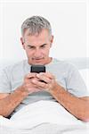 Smiling grey haired man sending a text in bed in bedroom at home
