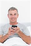 Happy grey haired man sending a text in bed looking at camera in bedroom at home
