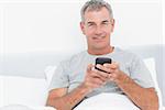 Cheerful grey haired man sending a text in bed smiling at camera in bedroom at home