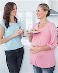 Smiling pregnant woman holding cookies and her friend in the kitchen