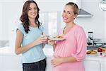Smling pregnant woman holding cookies and her friend standing in kitchen