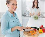 Blonde woman cutting carrots with her friend tossing salad in the kitchen