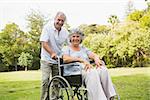 Mature woman in wheelchair with partner smiling at camera at the park