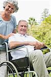 Cheerful mature man in wheelchair with partner looking at camera in park