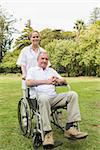 Happy man sitting in a wheelchair with his nurse pushing him smiling at camera in the park