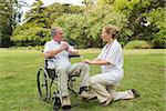 Smiling man in a wheelchair talking with his nurse kneeling beside him in the park on sunny day