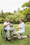 Happy man in a wheelchair talking with his nurse kneeling beside him on sunny day in the park
