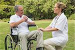 Cheerful man in a wheelchair talking with his nurse kneeling beside him in the park on a sunny day