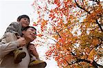 Little boy sitting on his fathers shoulders, walking through the park in autumn