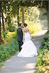 Backview of Bride and Groom kissing and holding hands, walking down pathway outdoors, on Wedding Day