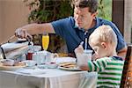 family of two eating nicely served breakfast outside; handsome young man pouring some coffee and his cute son eating delicious pancakes at breakfast time