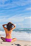 Rear view of beautiful young woman in bikini sitting cross legged on a deserted tropical beach with blue sky