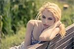 Portrait of beautiful blond lonely woman waiting on a wooden bench while resting her head on her arm.