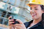 Young Professional Female Contractor Wearing Hard Hat at Contruction Site Texting with Cell Phone.