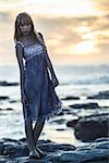 Beautiful model standing on rocks by the sea staring at camera at sunset