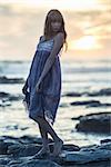 Beautiful model posing on rocks by the sea looking away at sunset