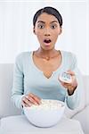 Surprised attractive woman sitting on cosy sofa eating popcorn while watching tv