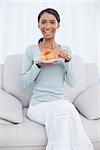 Smiling attractive woman eating croissant sitting on cosy sofa