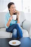 Happy woman holding cup of coffee eating cookie looking at camera