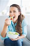 Happy attractive woman eating healthy salad sitting on cosy sofa