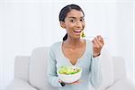 Smiling attractive woman sitting on cosy sofa in bright living room eating salad