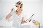 Peaceful cute blonde wearing hair curlers drinking coffee sitting on cosy bed