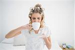 Peaceful blonde wearing hair curlers drinking coffee sitting on cosy bed
