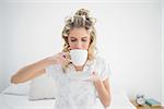 Relaxed pretty blonde wearing hair curlers drinking coffee sitting on cosy bed