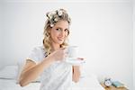 Relaxed pretty blonde wearing hair curlers holding coffee sitting on cosy bed
