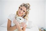 Smiling pretty blonde wearing hair curlers holding teddy bear sitting on cosy bed