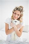 Peaceful pretty blonde wearing hair curlers holding teddy bear sitting on cosy bed