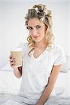 Peaceful pretty blonde wearing hair curlers holding coffee sitting on cosy bed