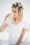 Peaceful pretty blonde wearing hair curlers drinking coffee sitting on cosy bed