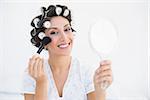 Smiling brunette in hair rollers holding hand mirror and applying makeup at home in bedroom