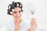 Happy brunette in hair rollers holding hand mirror and lip gloss at home in bedroom