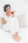Cheerful brunette in hair curlers sitting on her bed holding a cocktail in bedroom at home