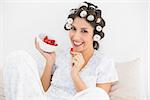 Cheerful brunette in hair rollers having a bowl of strawberries at home in bedroom