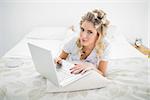 Serious pretty blonde wearing hair curlers using laptop lying on bed