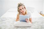 Thoughtful pretty blonde wearing hair curlers using tablet pc lying on cosy bed